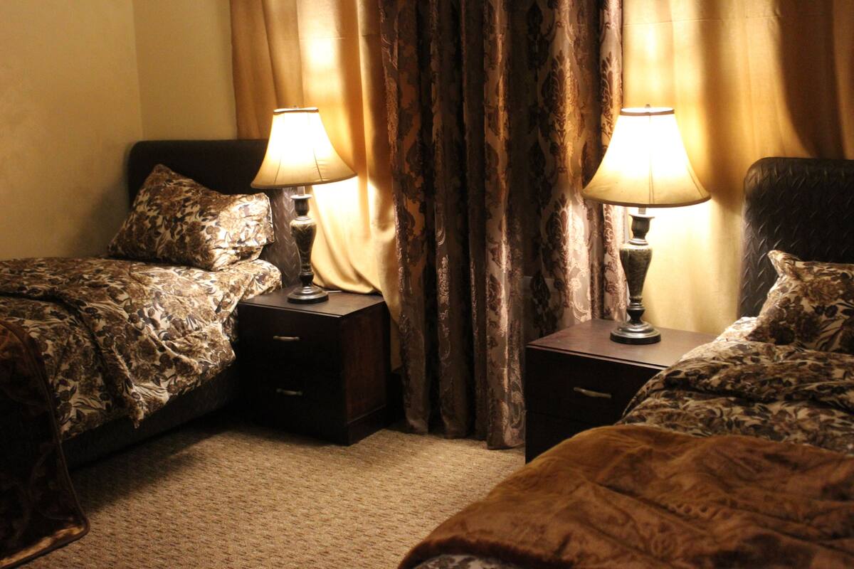 Une chambre d’hôtel avec deux lits, chacun recouvert d’une couvre-lit à motifs et agrémenté d’oreillers assortis. On y trouve deux tables de chevet avec des lampes, ainsi qu’une grande fenêtre avec des rideaux.