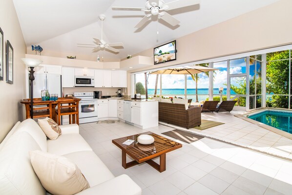 A modern kitchen with white cabinets, a dining area with a table and chairs, a living room with a sofa, and a poolside area with a view of the ocean.