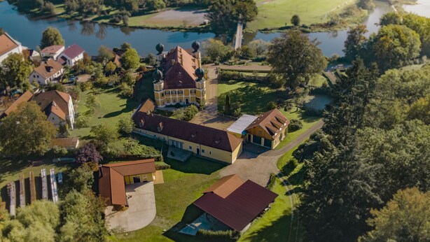 Luchtbeeld van een woongebied met huizen, een groot gebouw met een rood dak, en een waterpartij op de achtergrond.