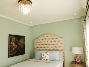 Une chambre à coucher avec une tête de lit capitonnée, un lit avec des oreillers, une œuvre d’art accrochée au mur, une lampe sur pied et un luminaire au plafond.