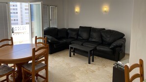 A living room with a black leather sofa, wooden dining table, and chairs. There are two wall-mounted lights and a sliding glass door leading to a balcony. - IFACH II 5th floor Gate 35 (Calp)