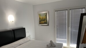 A neatly made bed with white linens and rolled towels, a black headboard, a framed painting on the wall, and a window with blinds. - IFACH II 5th floor Gate 35 (Calp)