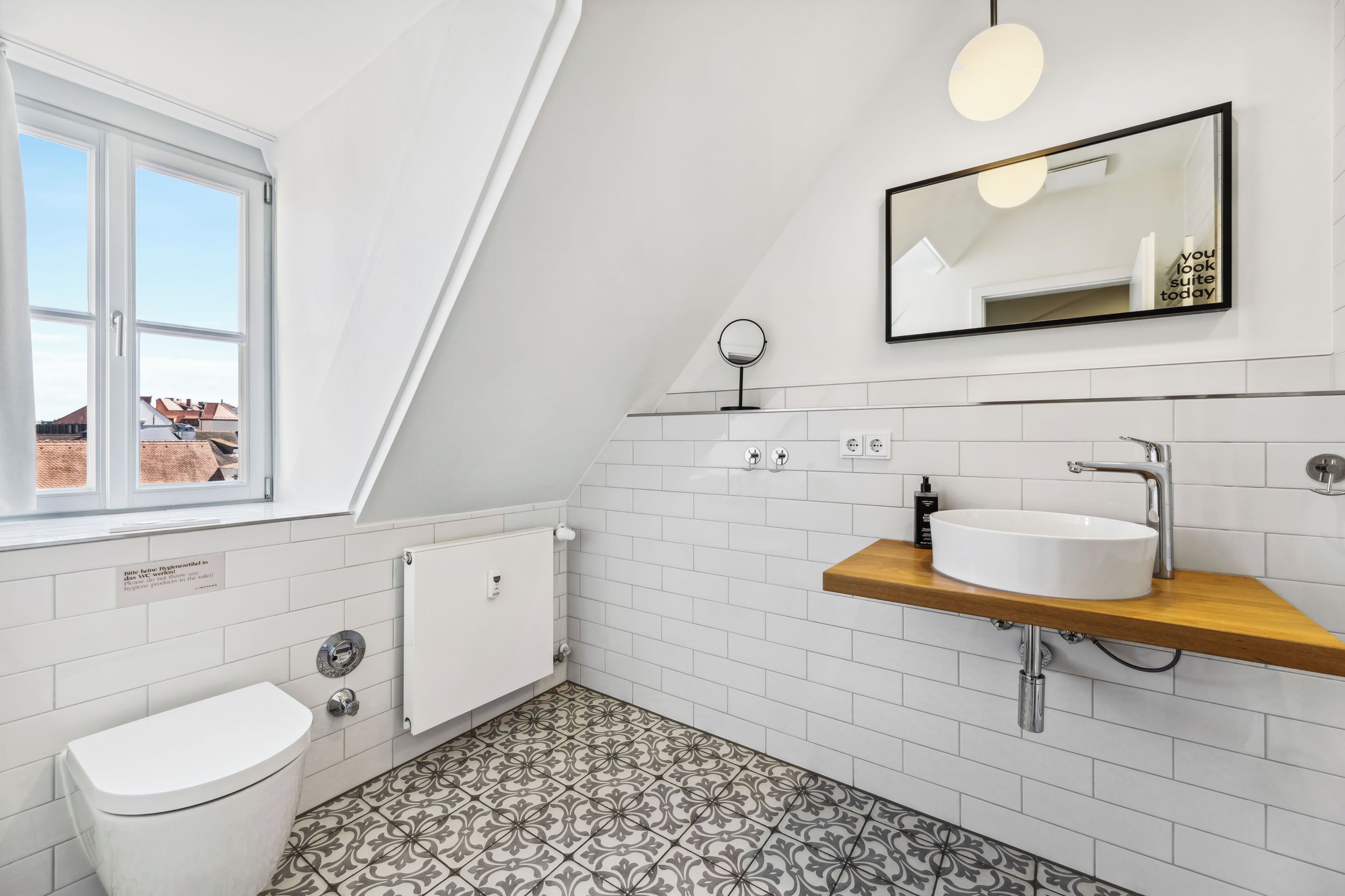 A modern bathroom with a white toilet, a sink with a wooden countertop, a mirror, and patterned floor tiles.