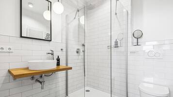 A modern bathroom with a white sink, wooden shelf, glass shower enclosure, and a white toilet.