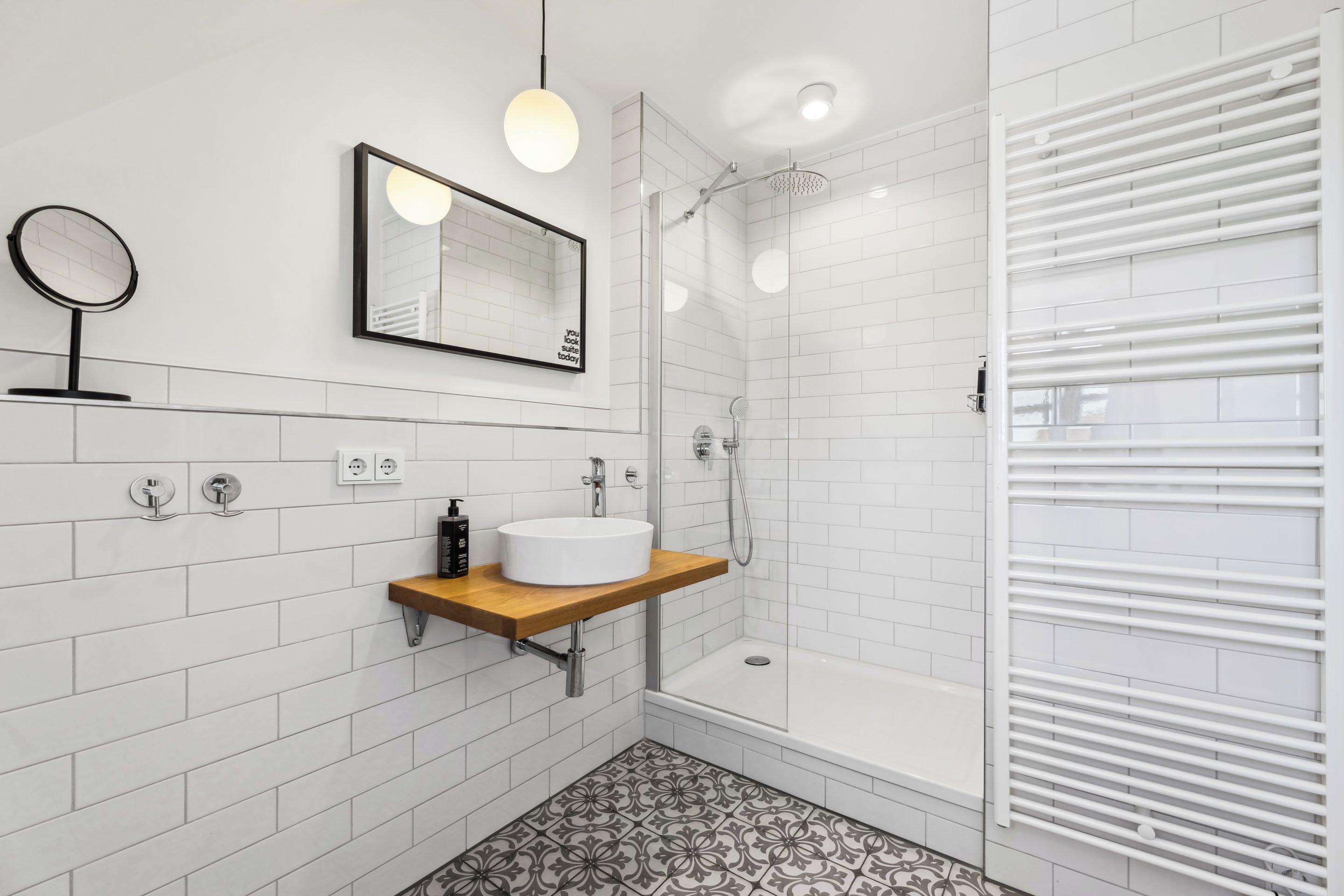 Un baño moderno con baldosas blancas tipo metro, un tocador de madera, un espejo redondo, un espejo grande, una ducha con puerta de vidrio y un radiador blanco.