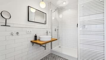 Un baño moderno con baldosas blancas tipo metro, un tocador de madera, un espejo redondo, un espejo grande, una ducha con puerta de vidrio y un radiador blanco.