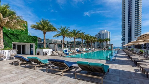 A rooftop pool area with lounge chairs, palm trees, and a view of the ocean and high-rise buildings.