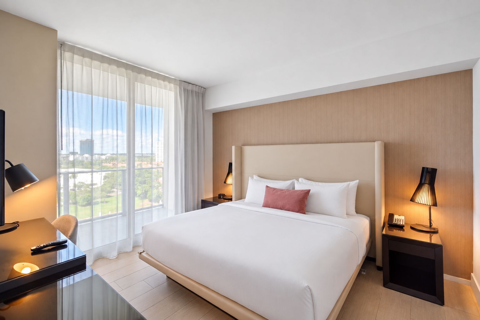 Habitación de hotel con una cama grande, un escritorio con lámpara, una silla y vista a la ciudad por la ventana.