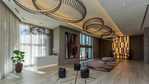 A spacious lobby with a modern design, featuring a large reception desk, multiple seating areas, and a large window providing natural light.
