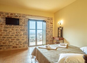 A hotel room with a stone wall, a bed with a beige bedspread, a wooden dresser, a flat-screen TV mounted on the wall, and a balcony with a view.