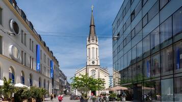 A plaza with a church steeple, potted plants, and people walking.