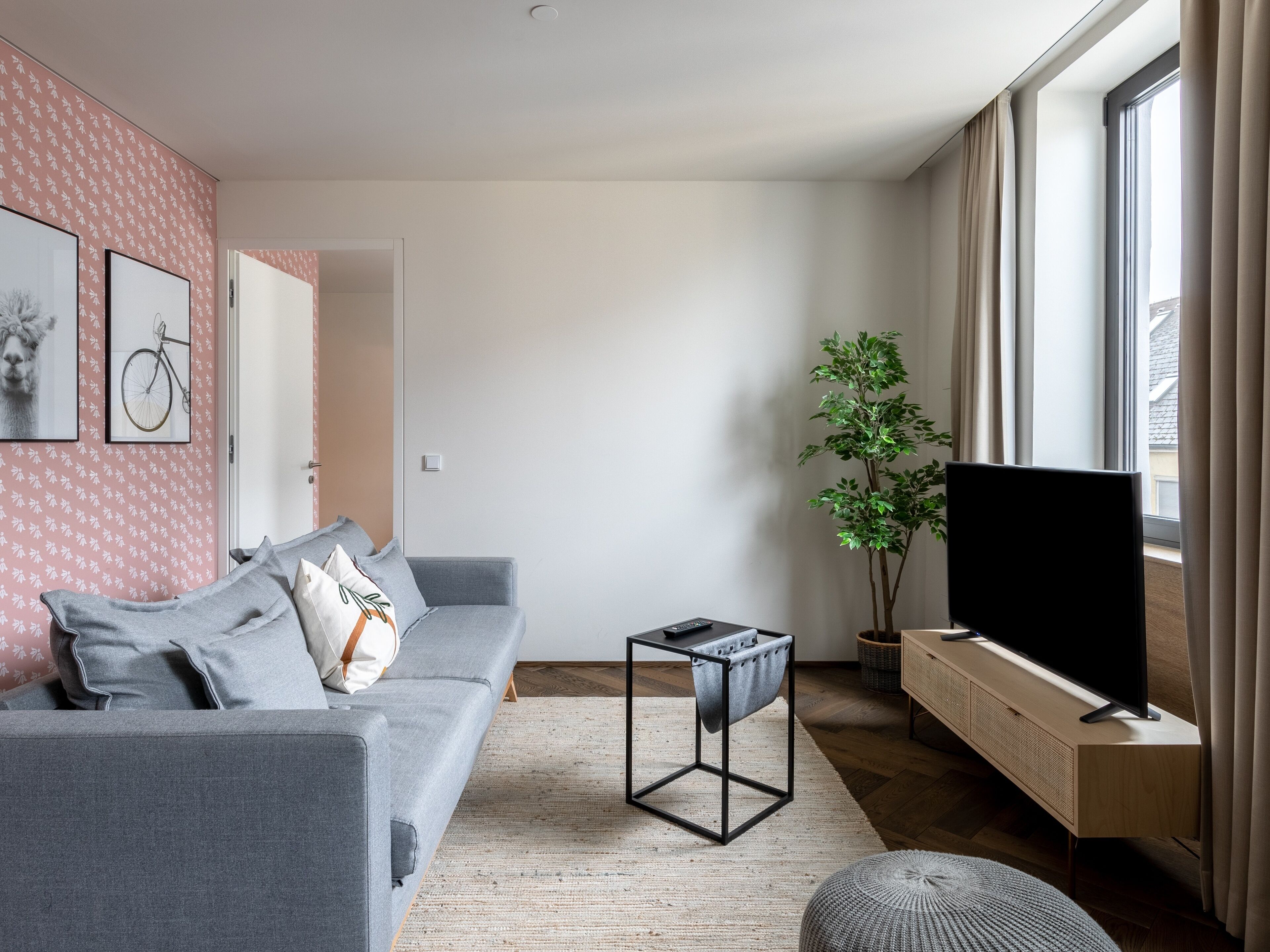 A modern living room with a grey sofa, a flat-screen TV, a potted plant, and a large window.