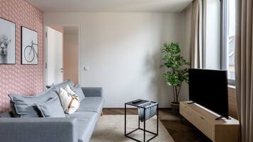 A modern living room with a grey sofa, a flat-screen TV, a potted plant, and a large window.