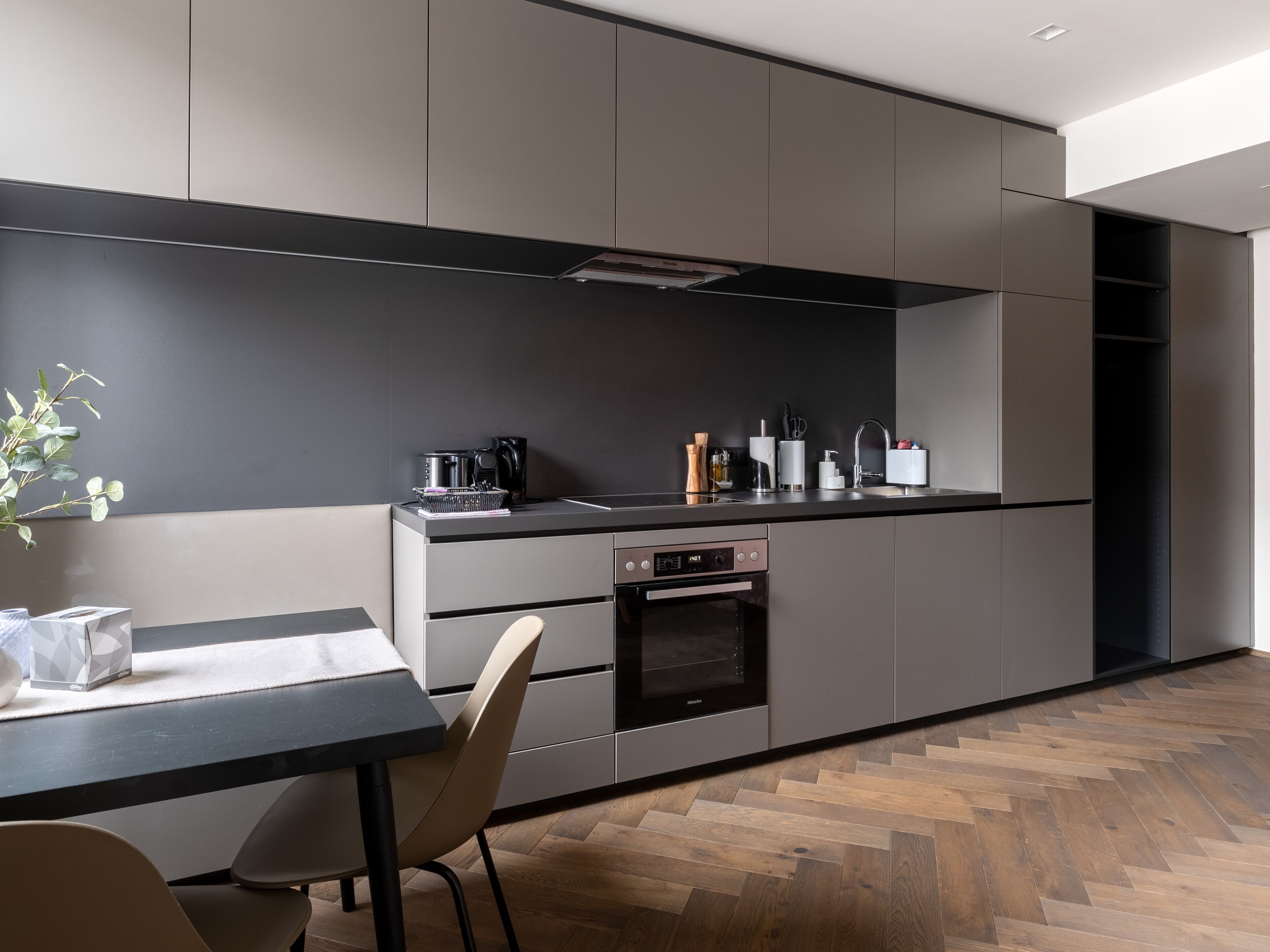 Una cocina moderna con salpicadero oscuro, electrodomésticos de acero inoxidable y suelo de madera en patrón de espiga.