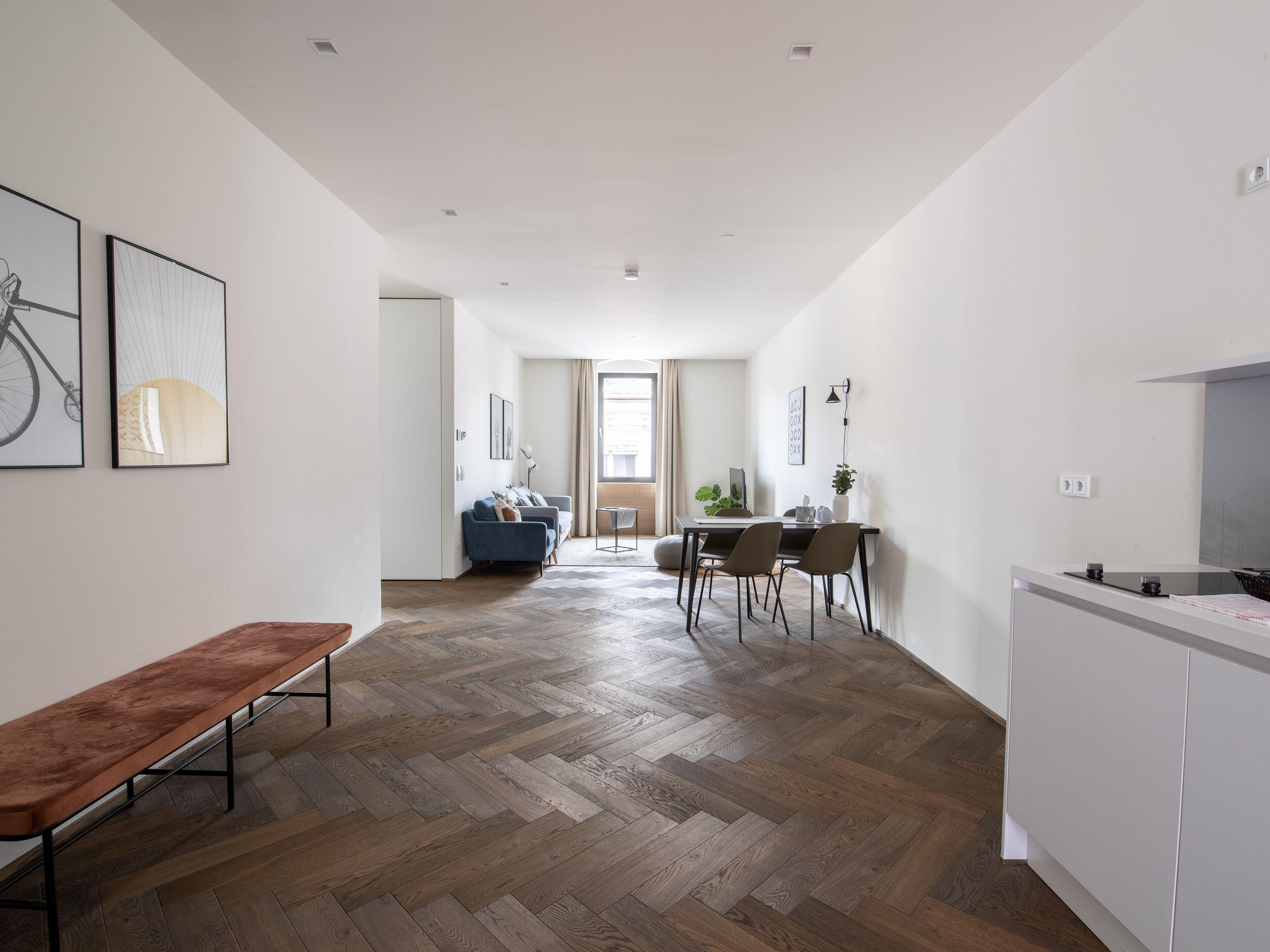 Un pasillo moderno y bien iluminado con piso de madera en patrón de espiga, un banco, un comedor con sillas y una mesa, y una kitchenette con estufa.