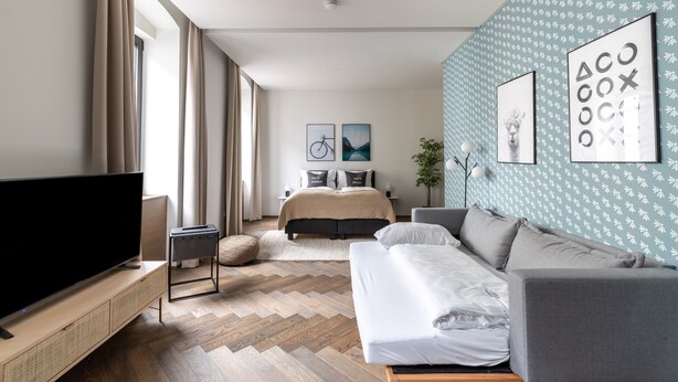 Una habitación de hotel moderna con un televisor grande, una cama, un sofá y un suelo con estampado tipo espiga.