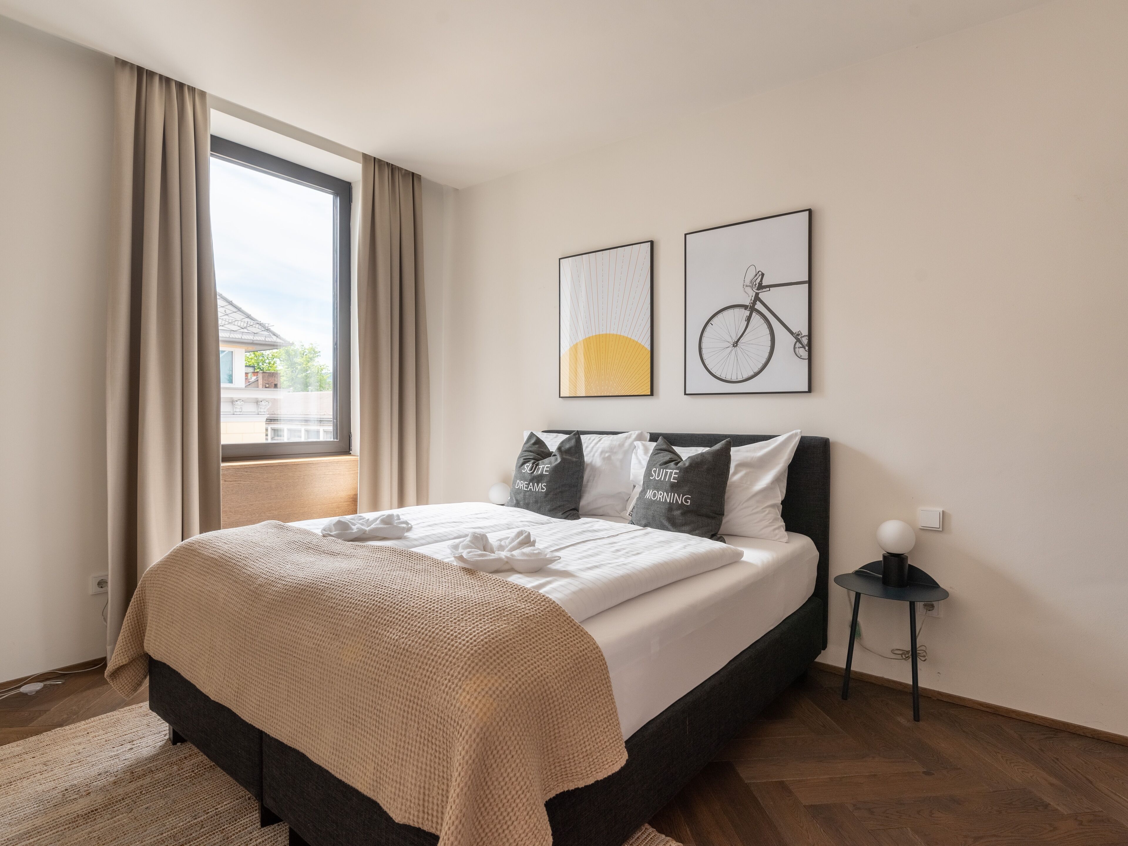 Una habitación de hotel moderna con una cama grande, una mesita de noche y un ventanal con cortinas. Hay dos cuadros en la pared.