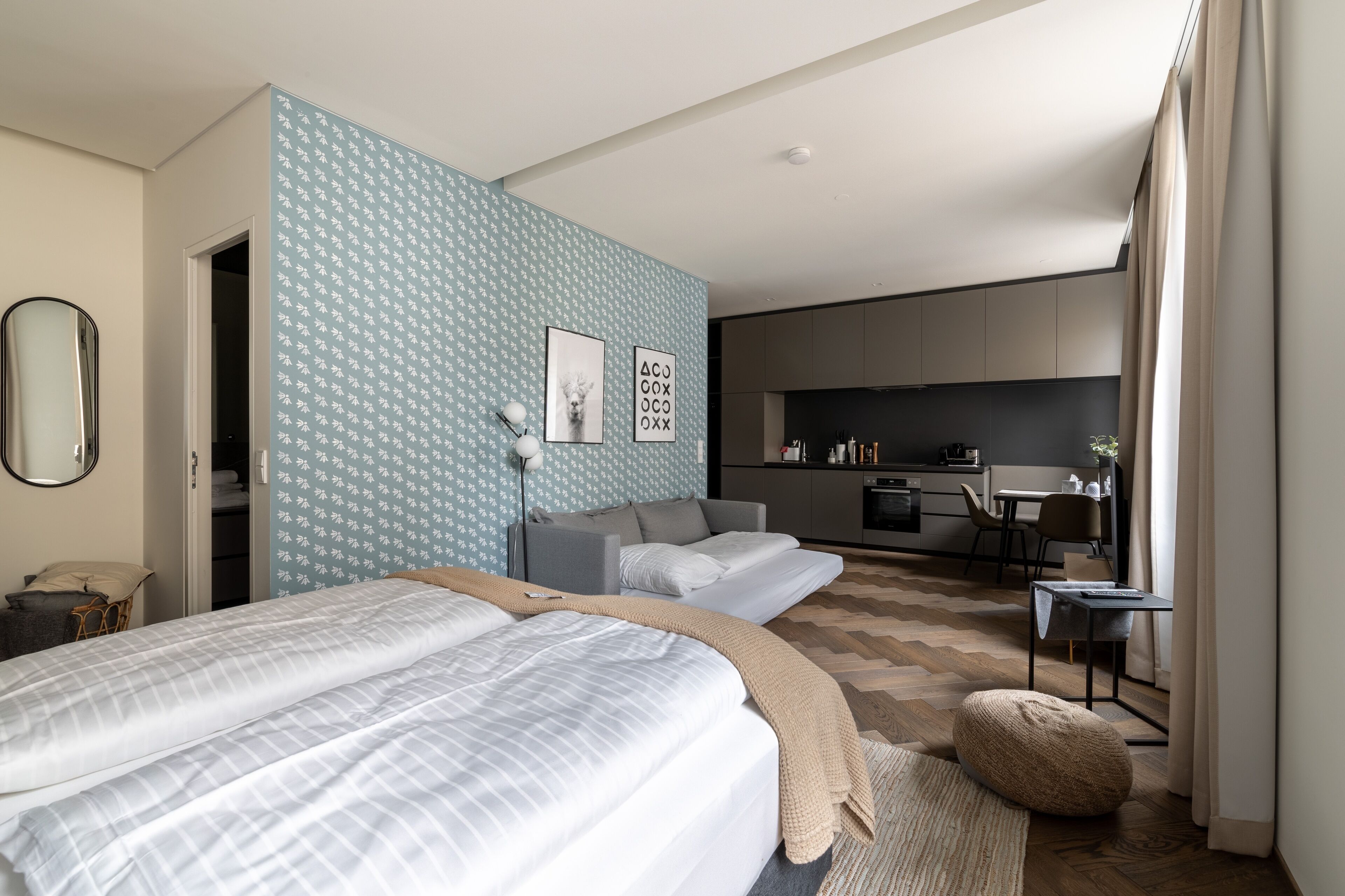 A modern bedroom with a large bed, a sofa, and a kitchenette in the background.