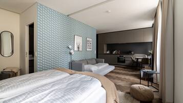 A modern bedroom with a large bed, a sofa, and a kitchenette in the background.