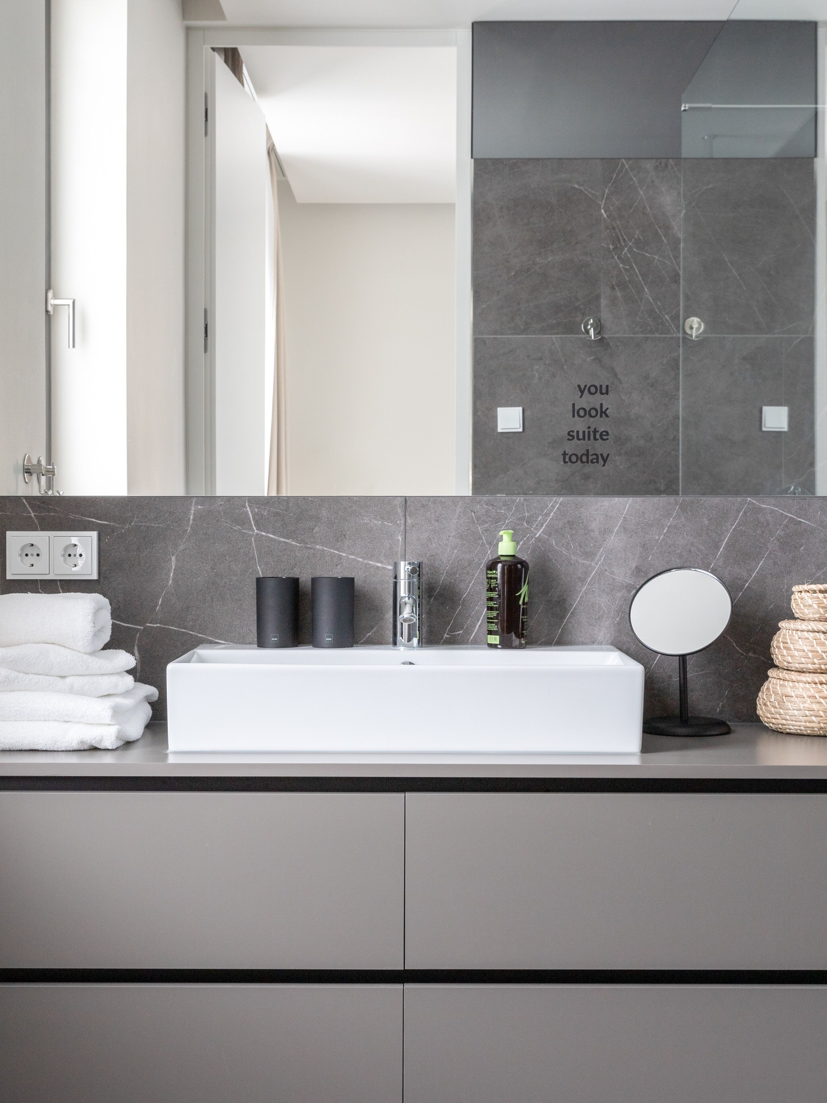 A modern bathroom with a white sink, black marble countertop, and a mirror. There are towels stacked on the counter, and a bottle of shampoo.