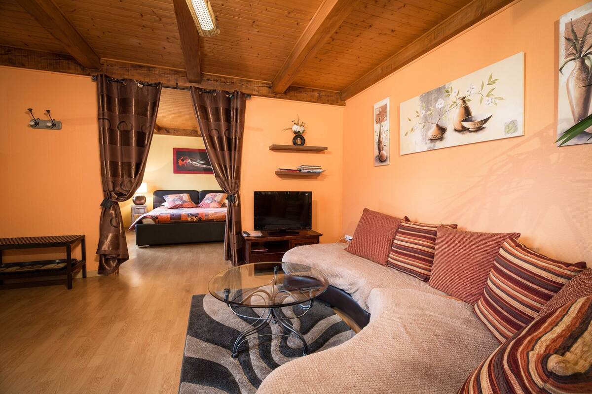 A cozy living room with a sofa, a glass coffee table, a TV, and a bed with a wooden headboard.