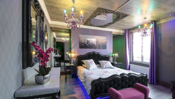 A bedroom with a large bed, a bedside table, a chair, a potted plant, and a chandelier. - Chambres et Gites La Paysanne (Fresnes-en-Woevre)