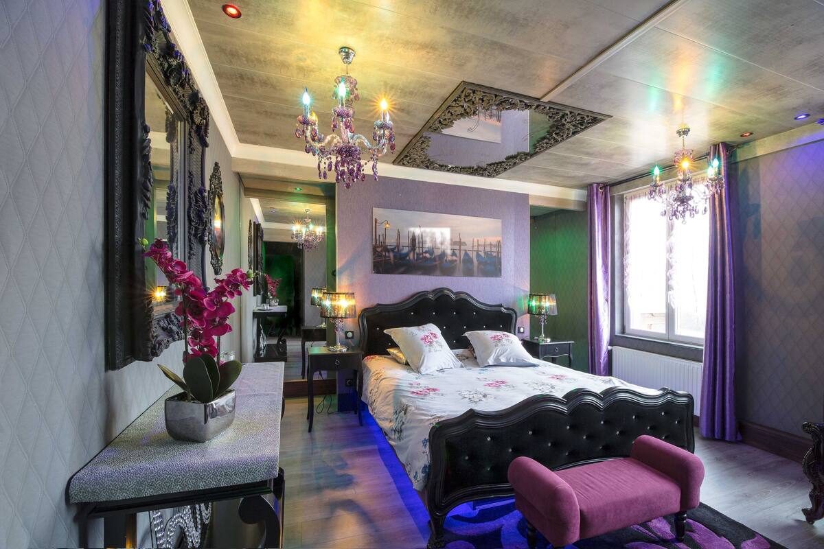 A bedroom with a large bed, a bedside table, a chair, a potted plant, and a chandelier.