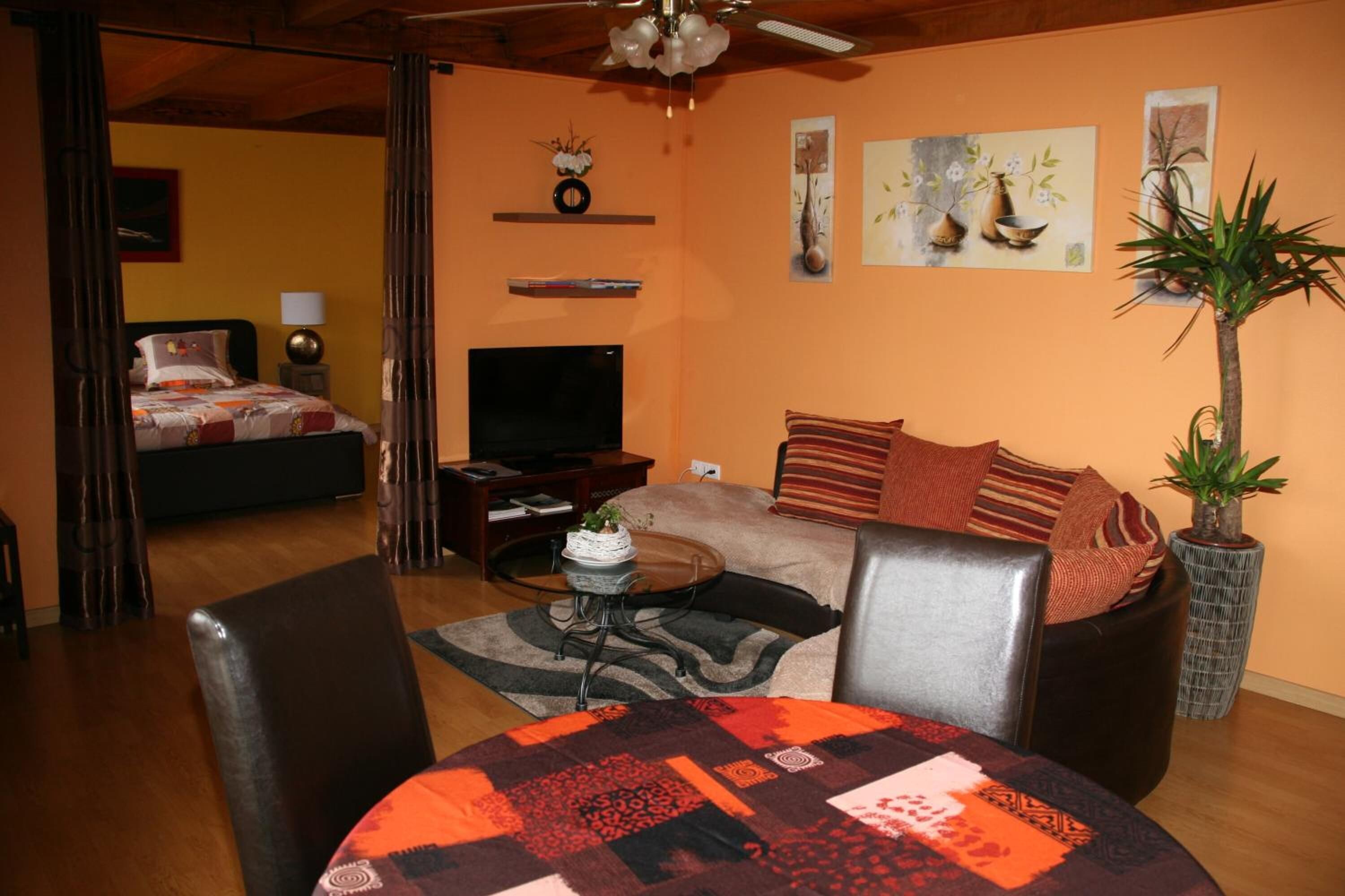 A cozy living room with a sofa, a coffee table, and a TV. There is a bed in the background and a plant on the right.