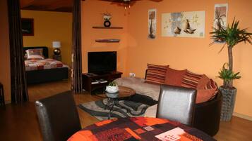 A cozy living room with a sofa, a coffee table, and a TV. There is a bed in the background and a plant on the right.