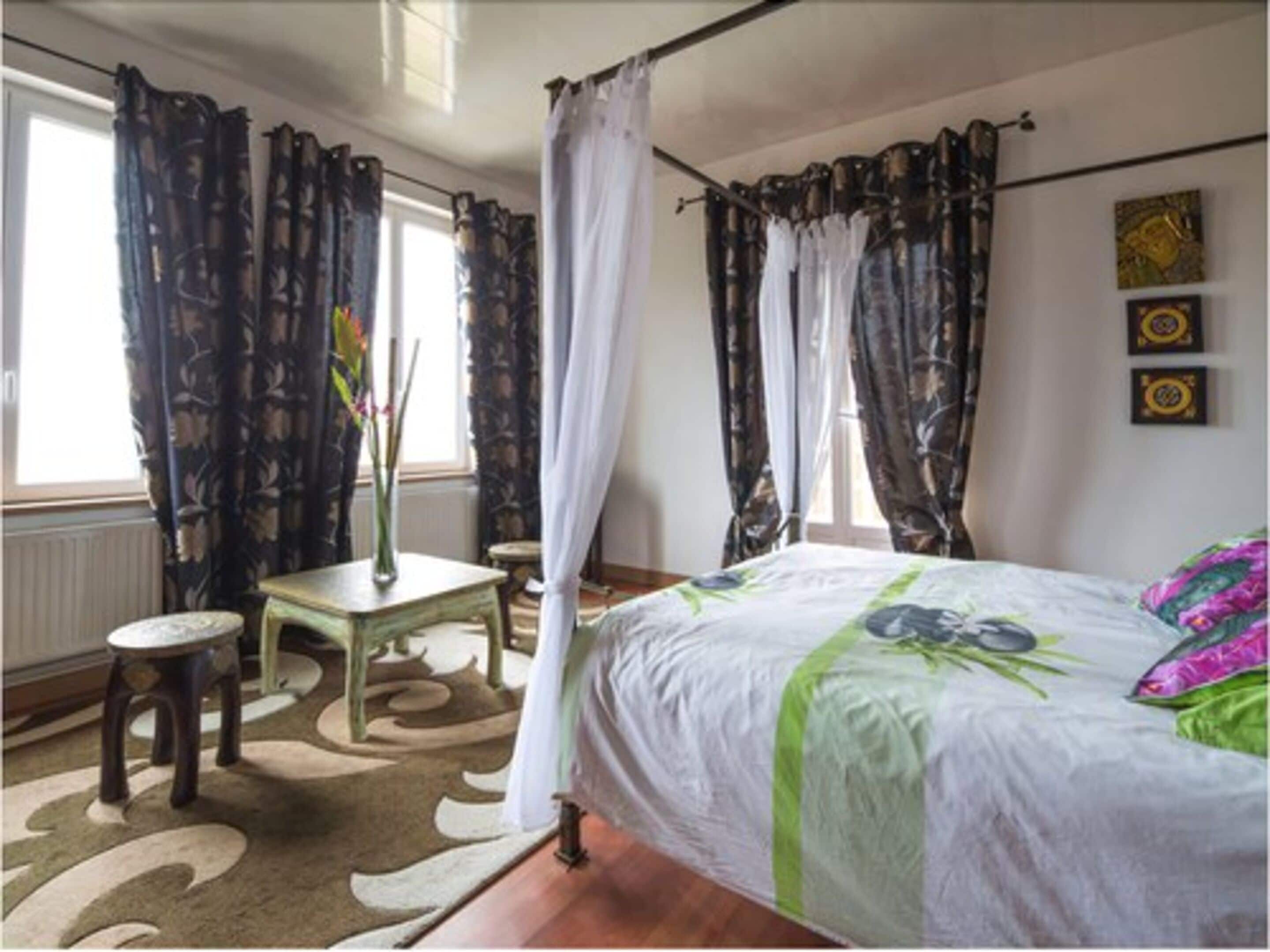 Un dormitorio con una cama, una mesita, una silla y un ventanal con cortinas.