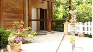A wooden building with a porch, potted plants, and a white picket fence. - Chambres et Gites La Paysanne (Fresnes-en-Woevre)