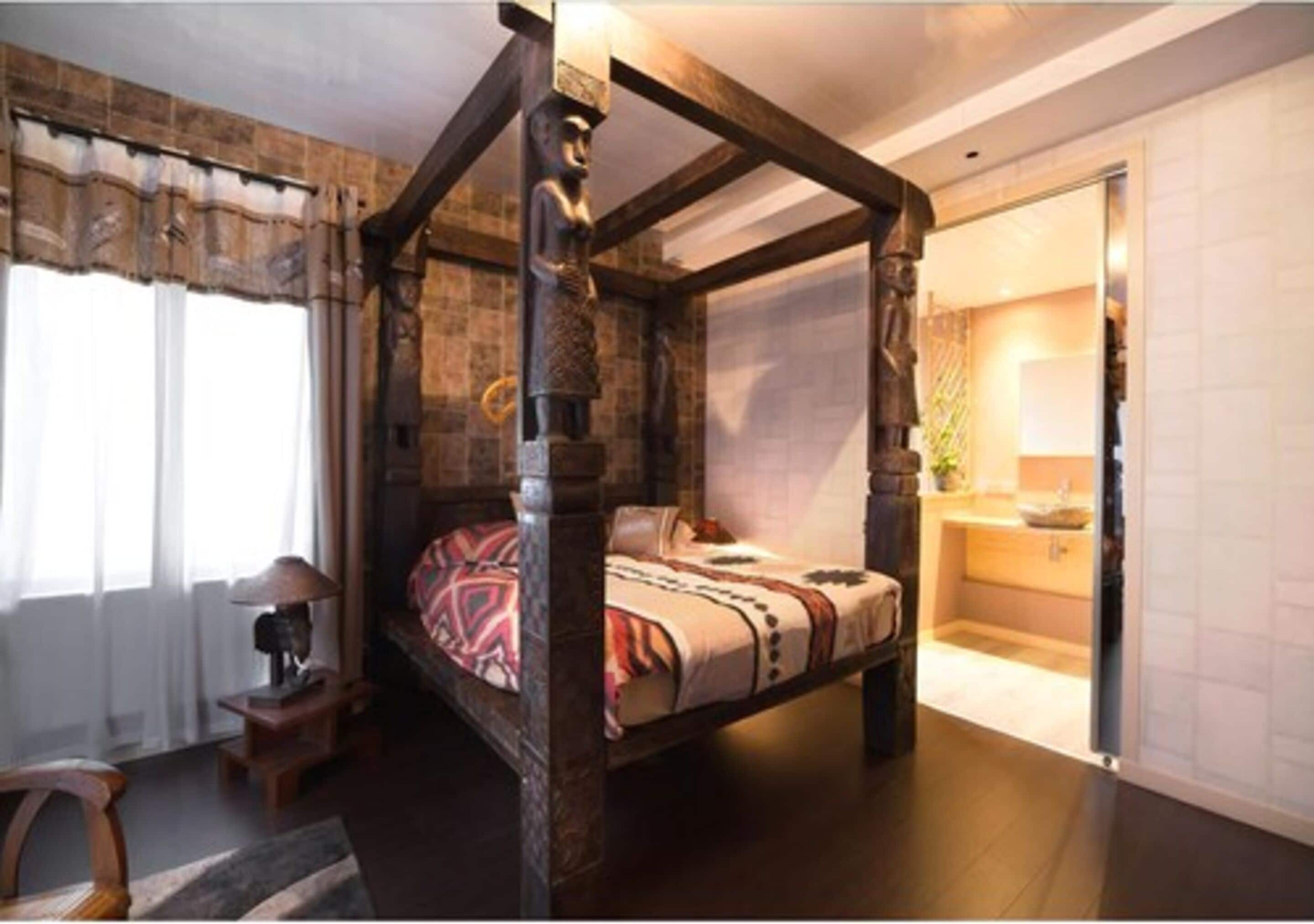 A four-poster bed with a patterned bedspread, a small bedside table with a lamp, and a view of a bathroom with a sink and mirror.