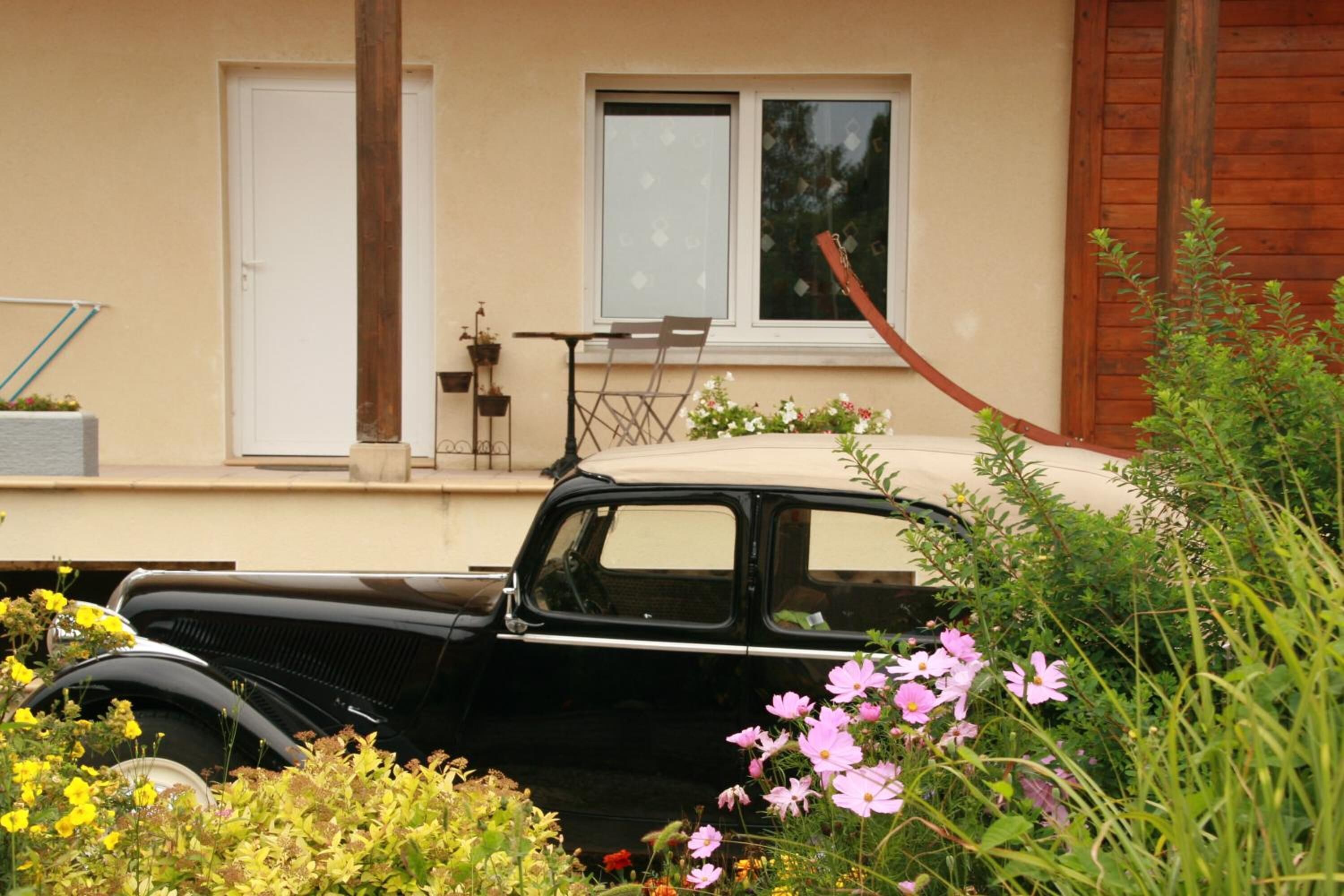 A vintage black car parked in front of a house with a window, a door, and a small table with chairs.