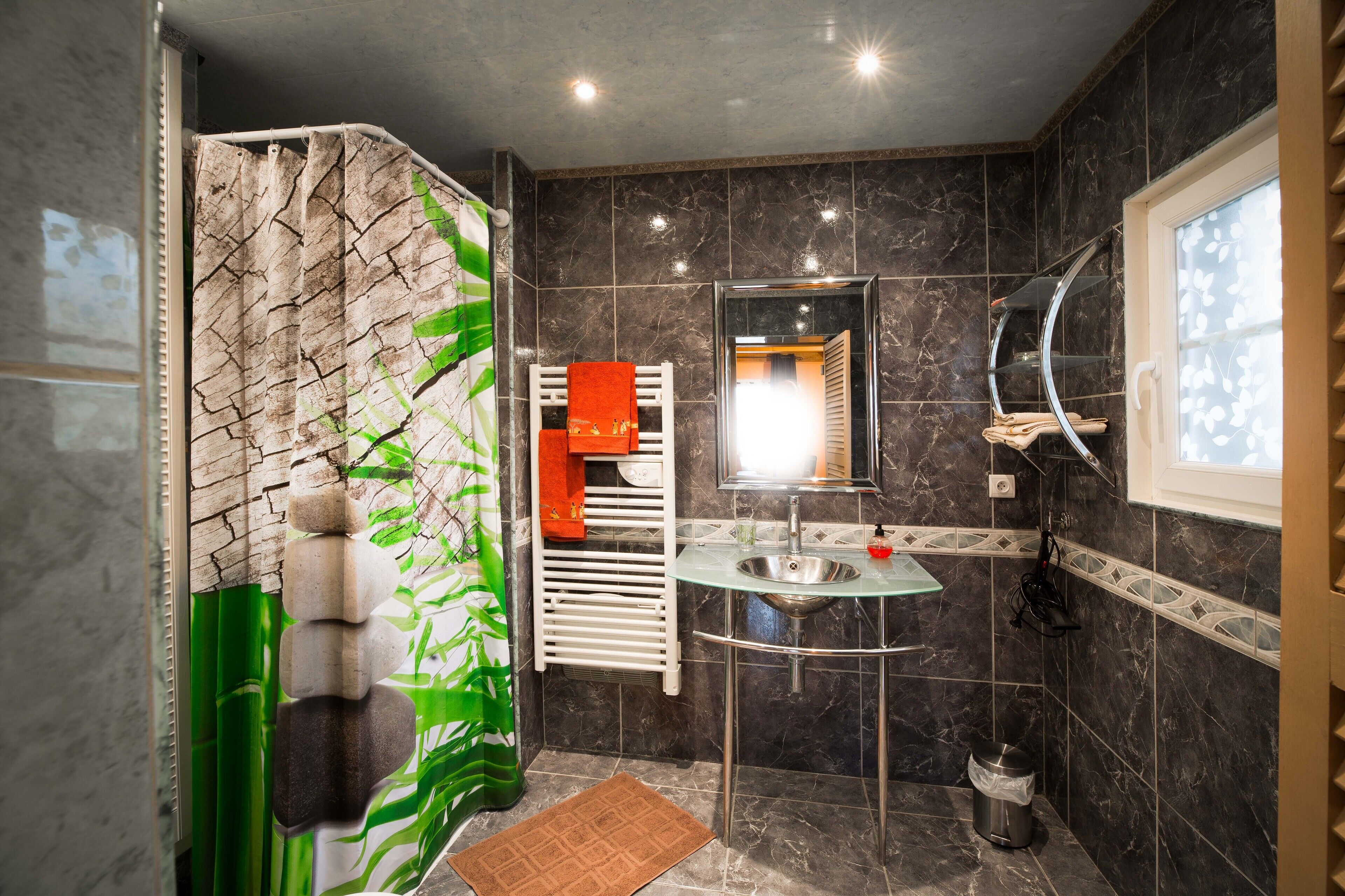 Une salle de bain moderne avec un lavabo en verre, un porte-serviettes blanc, un miroir et un rideau de douche orné d’un motif de feuilles vertes.