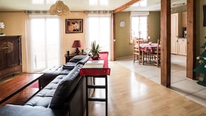 A spacious living room with a sofa, a dining table, and a kitchen area visible through an open doorway. - Chambres et Gites La Paysanne (Fresnes-en-Woevre)
