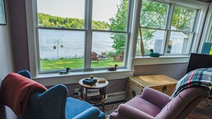 A cozy room with a large window overlooking a lake, featuring two chairs, a small table, and a wicker basket.