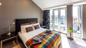 Ein modernes Hotelzimmer mit einem großen Bett, einem Nachttisch, einer Lampe und Blick auf die Stadt durch große Fenster.