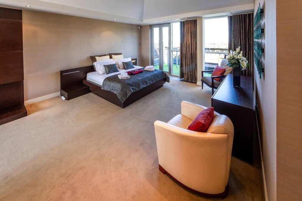 A modern hotel room with a large bed, a seating area with a chair, and a view of the outdoors through a sliding door.