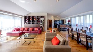 Ein großzügiger Wohnbereich mit einem roten Sofa, einem beigen Sofa und einem Esstisch mit Stühlen. Ein Bücherregal ist mit Büchern und Dekoration gefüllt.