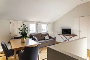A modern living room with a sofa, a dining table, chairs, a TV on a stand, and a large window with curtains. - Citybreak Apartments Lapa - Budget Duplex Studio (Porto)