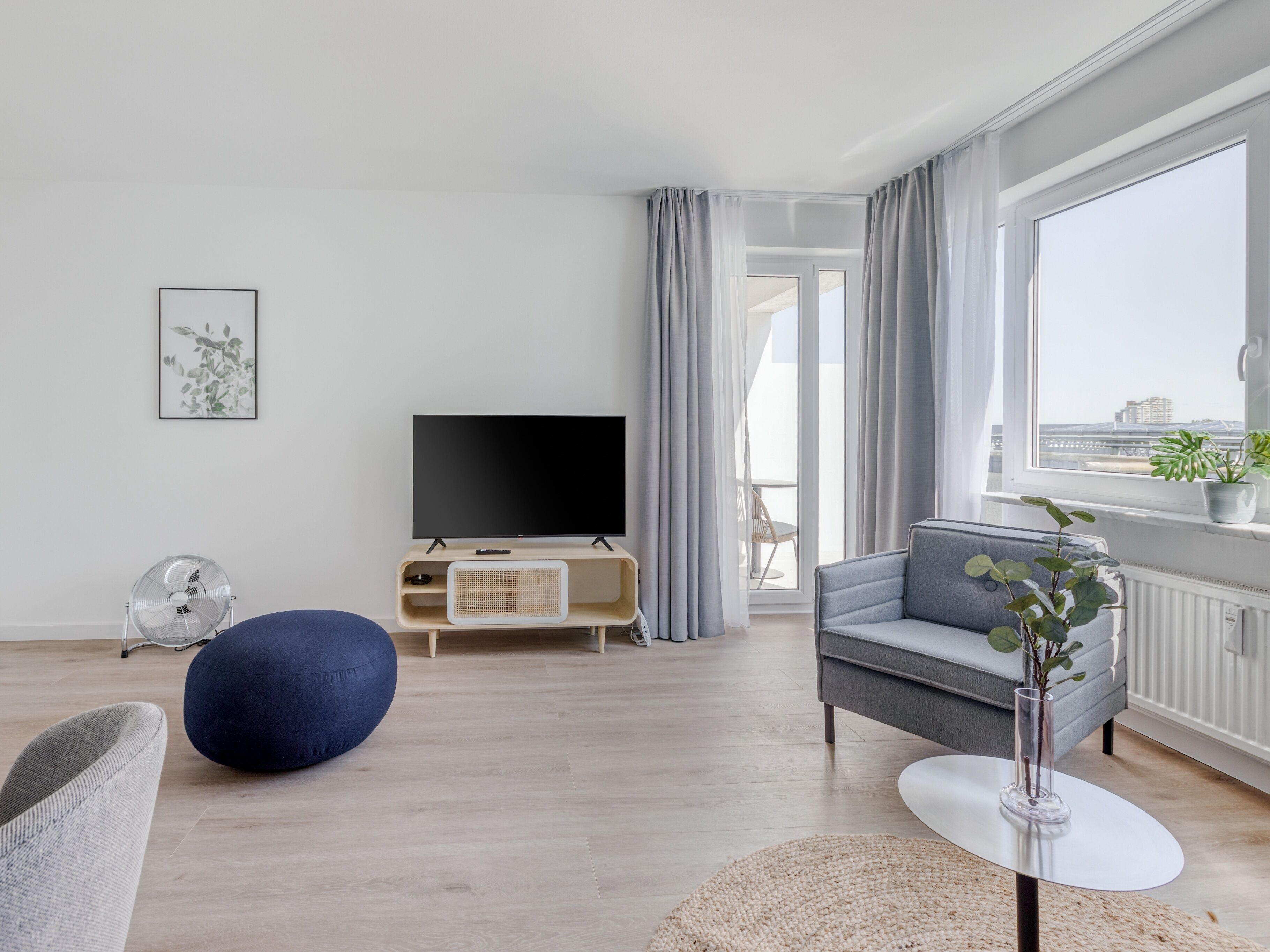 Una sala de estar moderna con un televisor de pantalla plana, un sofá gris, un sillón de pelotas azules y vistas a la ciudad a través de amplias ventanas.