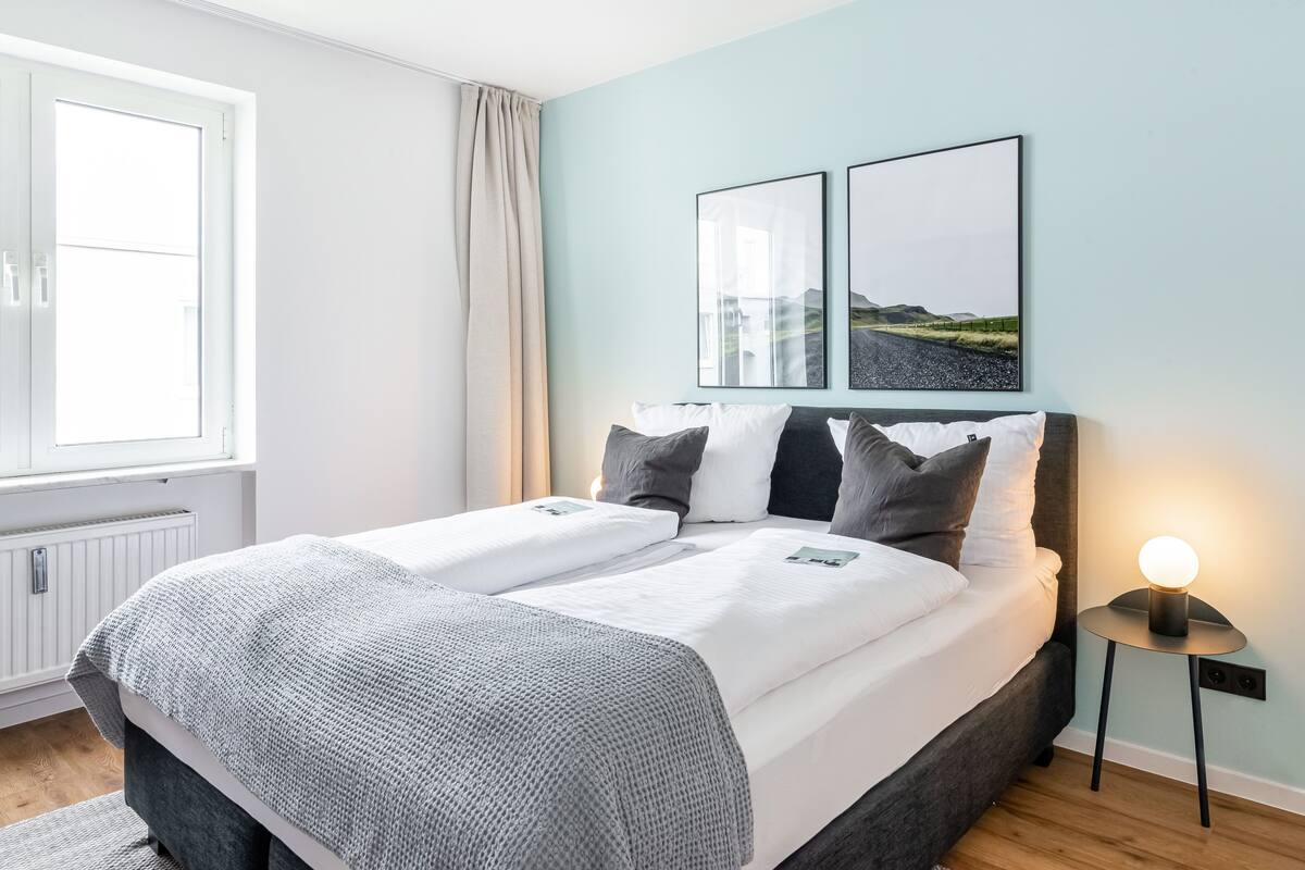 Ein ordentlich bezogenes Doppelbett mit einer grau-weißen Decke, ein Nachttisch mit Lampe und zwei gerahmte Bilder an der Wand.