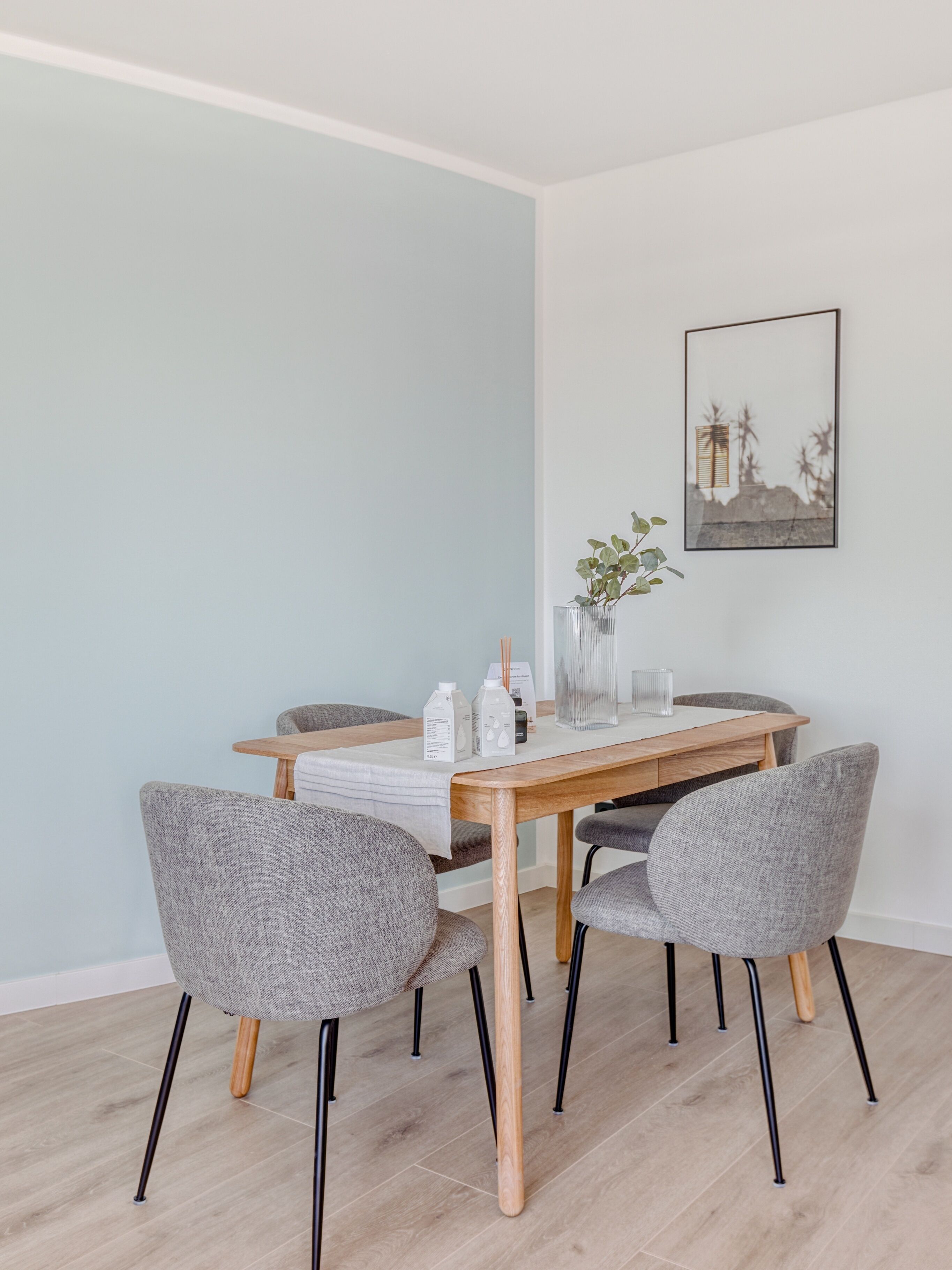 A dining area with a wooden table, four grey upholstered chairs, a white tablecloth, and a framed picture on the wall.