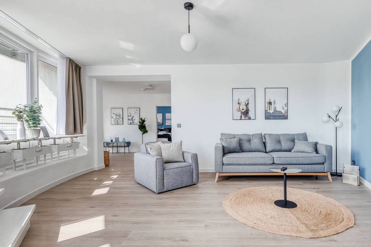 Ein modernes Wohnzimmer mit einem grauen Sofa, einem kleinen grauen Sessel und einem gewebten Teppich. Im Hintergrund befindet sich ein Essbereich mit Tisch und Stühlen.