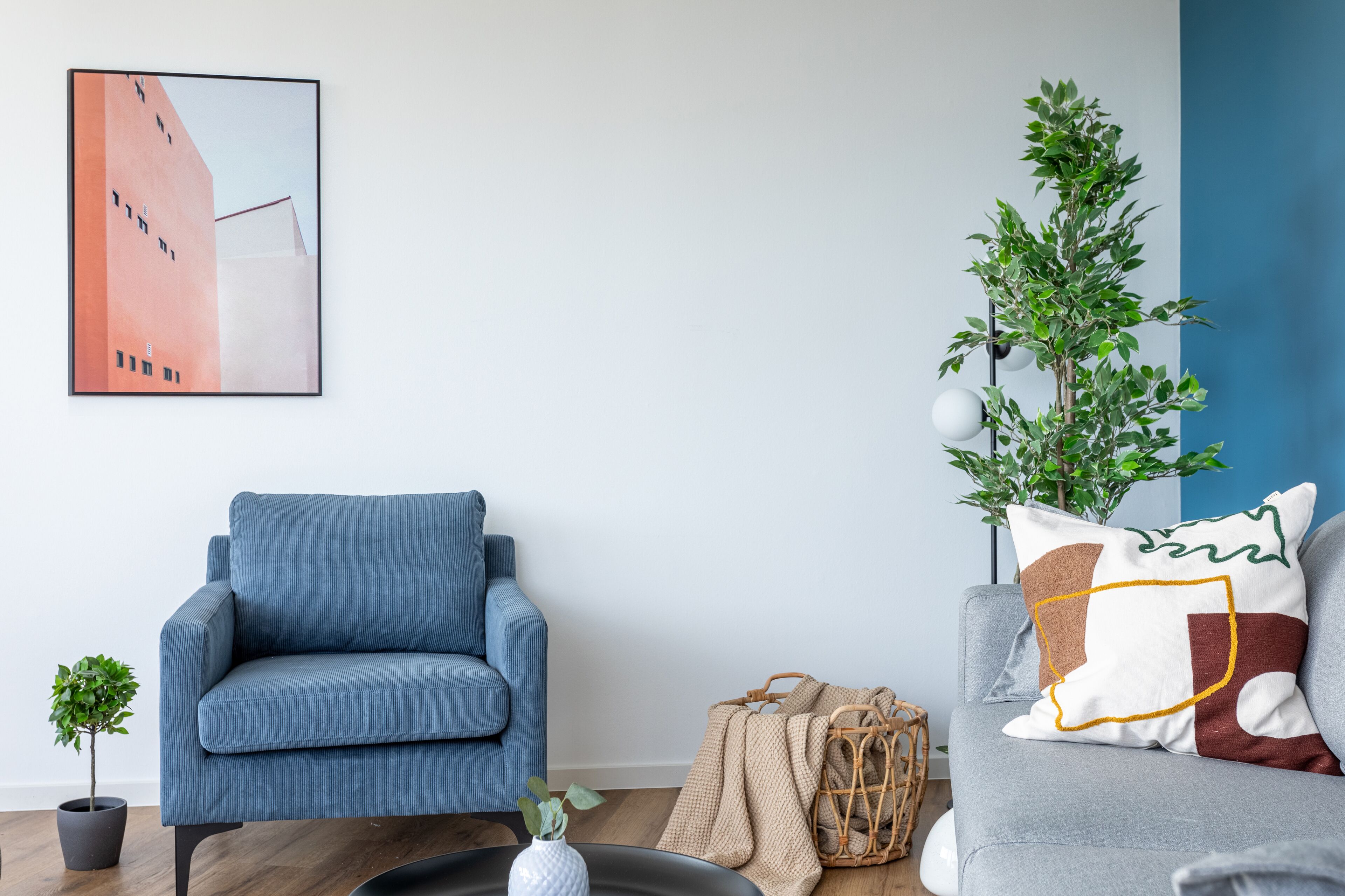 Una sala moderna con un sillón azul, un sofá gris, una pequeña planta en maceta, una cesta de mimbre y un cuadro en la pared.