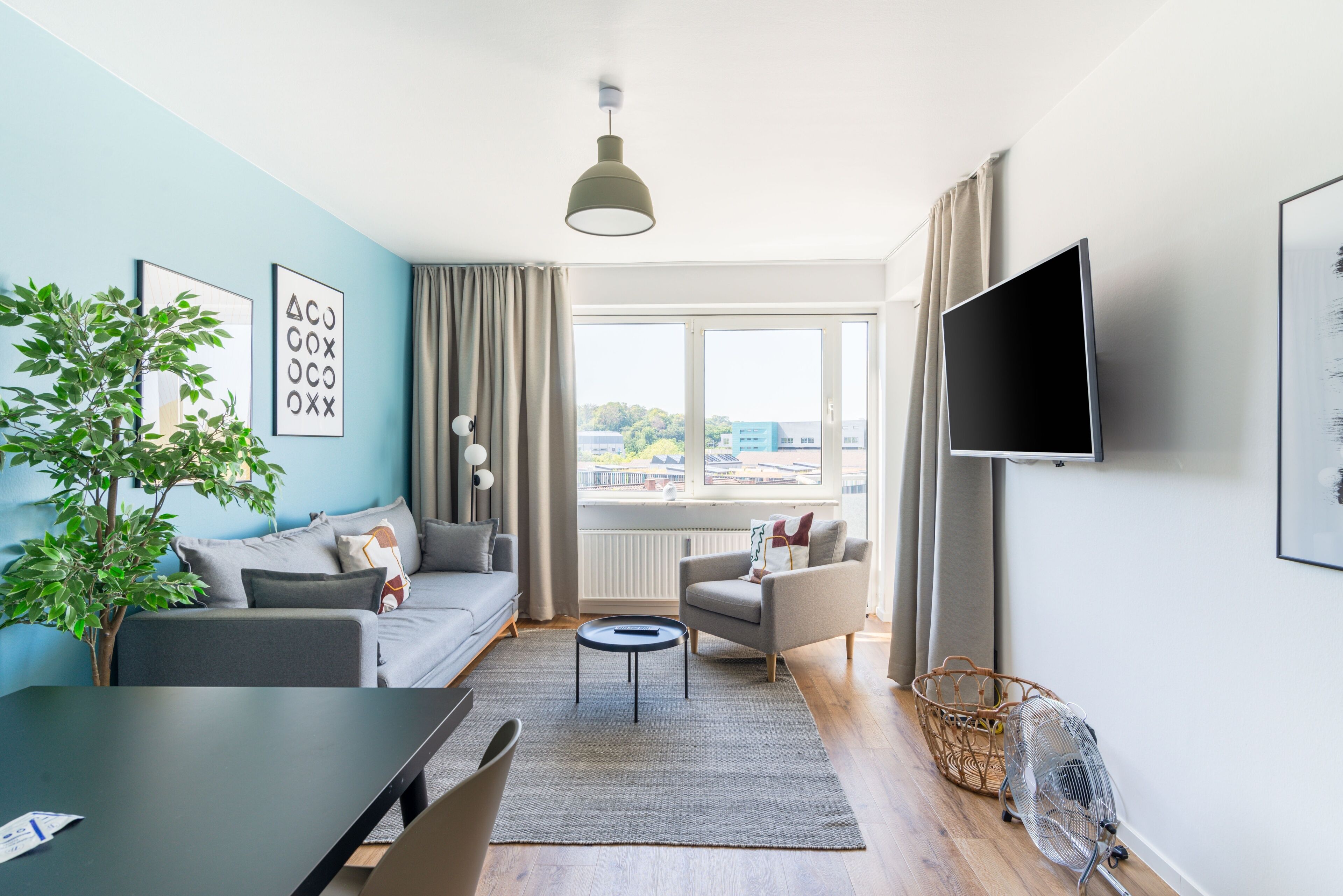 Una sala moderna con sofá, sillón y mesa de comedor. Hay un televisor montado en la pared y un ventanal con vistas a la ciudad.