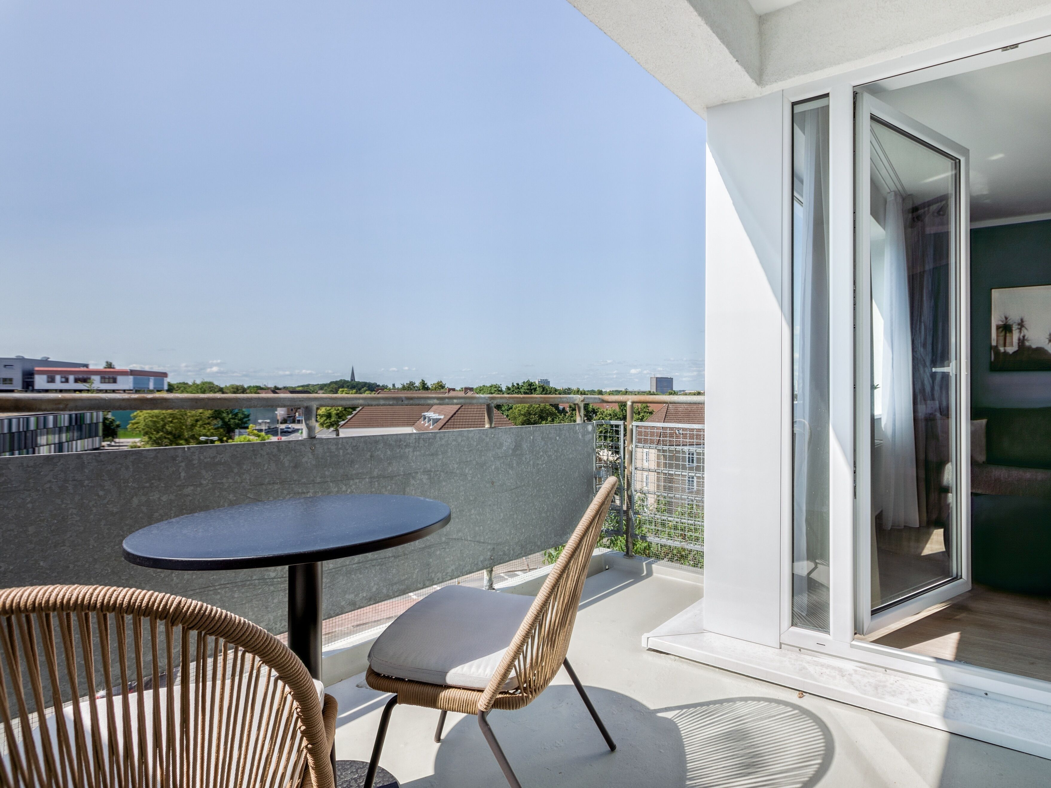 A balcony with a round table and wicker chair, overlooking a cityscape with buildings and greenery.