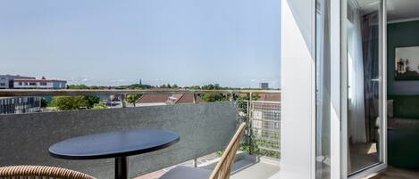 A balcony with a round table and wicker chair, overlooking a cityscape with buildings and greenery.