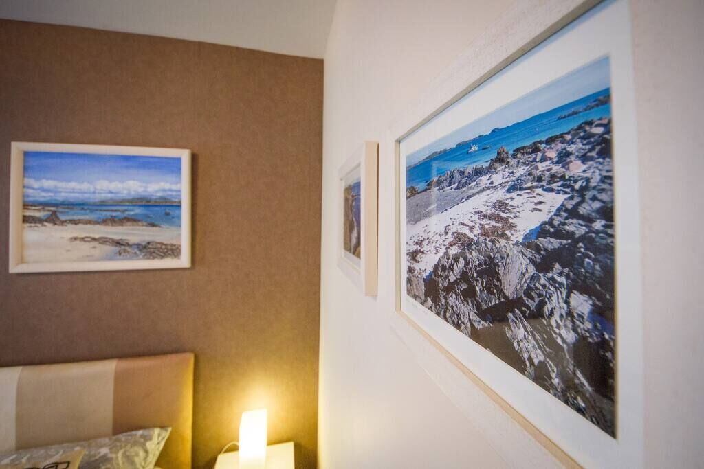A room with a bed, a wall-mounted lamp, and two framed photographs depicting coastal landscapes.