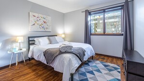 Un dormitorio ordenado con cama, lámparas de noche, una ventana con cortinas, una obra de arte en la pared y una alfombra estampada.
